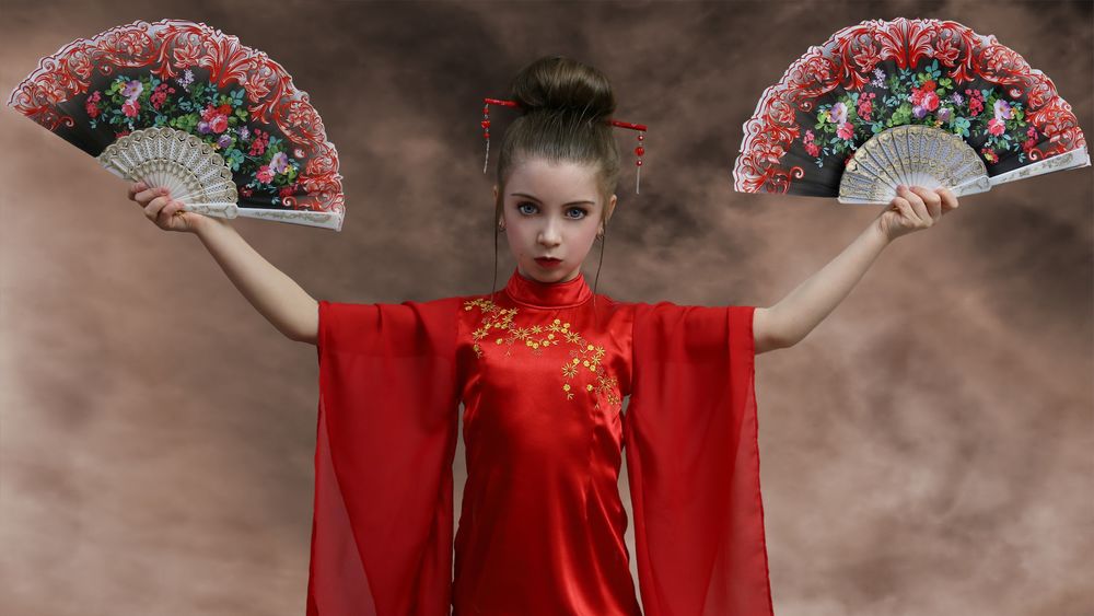 Обои для рабочего стола Восточная девочка в красном платье стоит с серьезным выражением лица с веерами в обеих руках