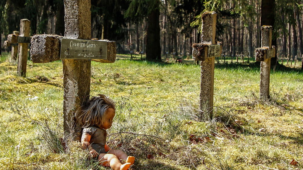 Обои для рабочего стола На старом заброшенном кладбище под замшелым, поросшем мхом, крестом лежит кукла