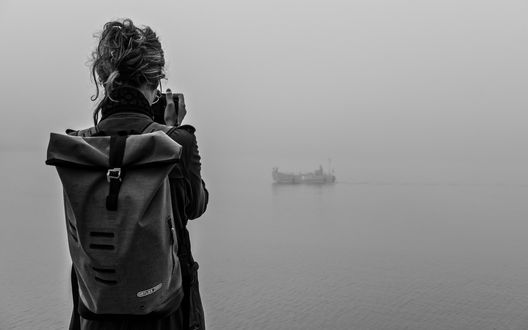 Обои на рабочий стол Девушка с рюкзаком за спиной фотографирует лодку посреди туманного озера