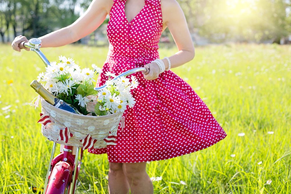Обои для рабочего стола Девушка в платье в горошек стоит с велосипедом, на котором корзина с ромашками и бутылкой шампанского