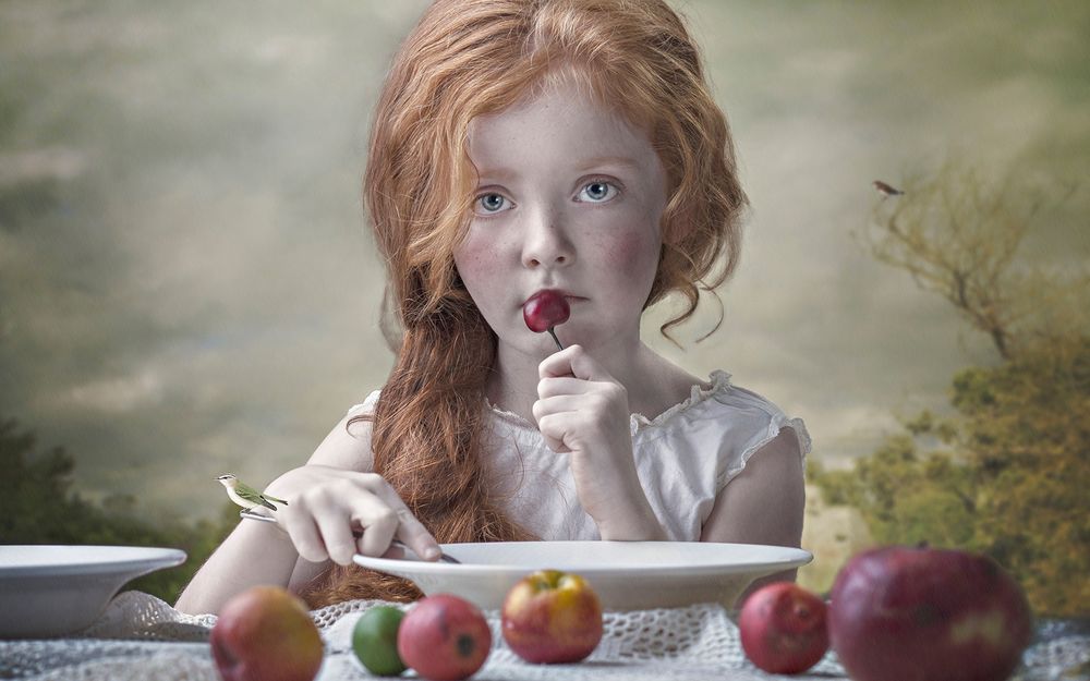 Обои для рабочего стола Рыжеволосая девочка сидит перед тарелкой за столом, с вишней в руке, которую она держит к рта, by Ewa Cwikla