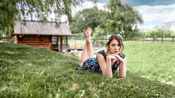 Обои на рабочий стол Модель Disha Shemetova / Диша Шеметова в легком летнем платье позирует, лежа на траве, на размытом фоне деревьев и деревяного дома