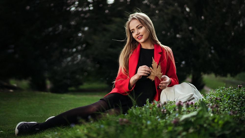 Обои для рабочего стола Модель Maria Puchnina / Мария Пучнина в красном пальто позирует, сидя на лужайке на размытом фоне деревьев. Фотограф Иван Горохов