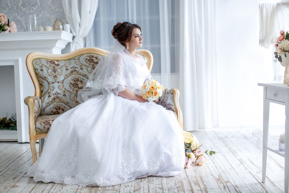 Обои для рабочего стола Невеста в свадебном платье с букетом сидит на диване в светлой комнате с камином