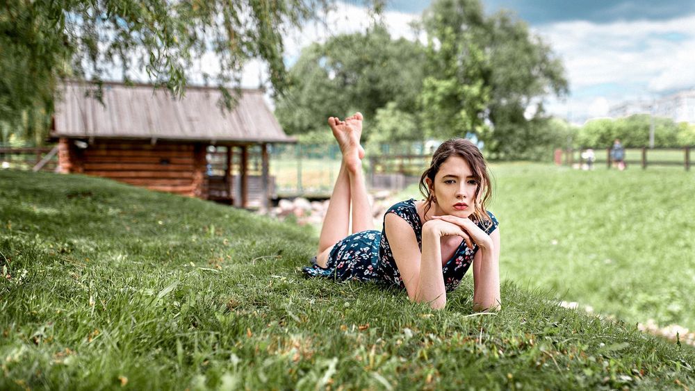Обои для рабочего стола Модель Disha Shemetova / Диша Шеметова в легком летнем платье позирует, лежа на траве, на размытом фоне деревьев и деревяного дома