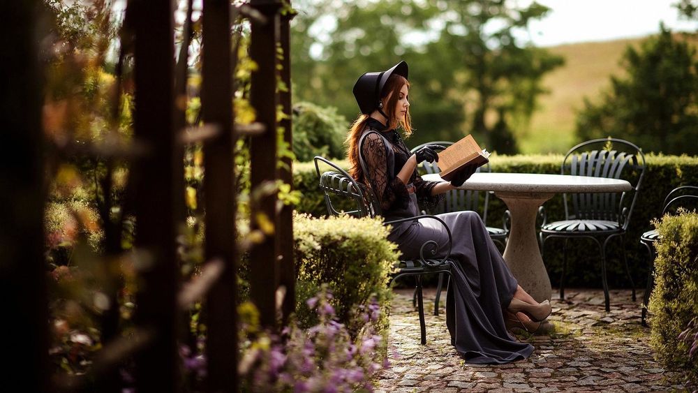 Обои для рабочего стола Модель Виктория Марчук в шляпке, черной кружевной блузке и в длинном сарафане позирует, сидя за столом в саду с книгой в руках. Фотограф Елена Смирнова