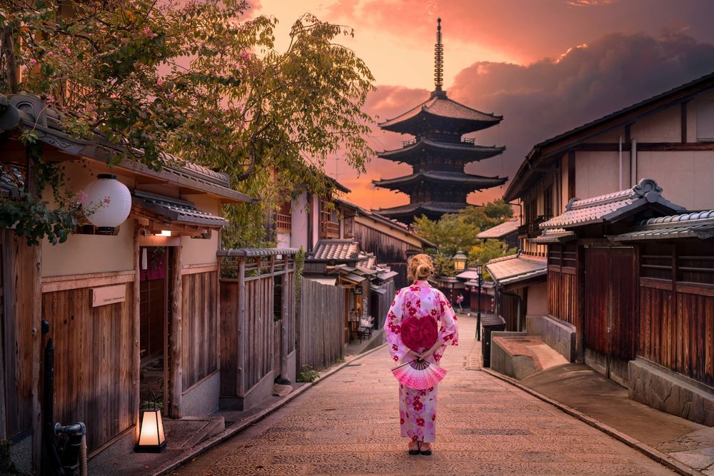Обои для рабочего стола Японская девушка в кимоно с веером стоит спиной на старой улочке с видом на пагоду, Киото / Kyoto, Япония / Japan