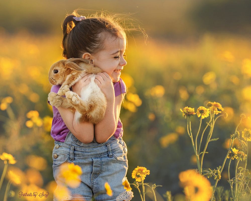 Обои для рабочего стола Девочка с радостью и нежностью прижимает к себе кролика, стоя в цветущем поле, фотограф Suzy Mead