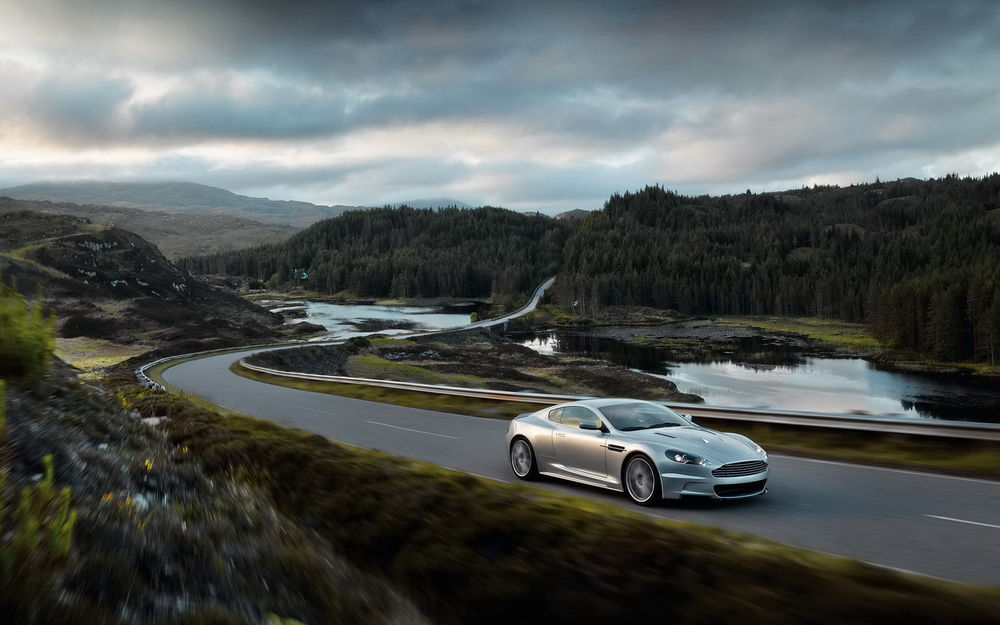 Обои для рабочего стола Серебристый Aston Martin DBS едет на фоне холмов покрытых лесом