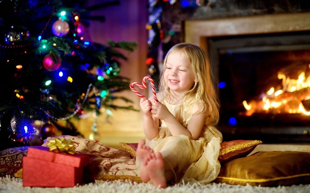 Обои для рабочего стола Радостная девочка сидит на подушках у новогодней елки и камина