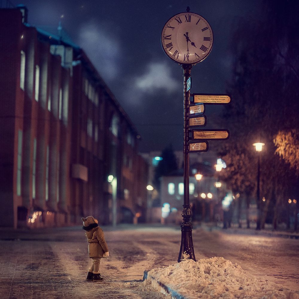 Обои для рабочего стола Ребенок в зимней одежде стоит на улице вечернего города и смотрит на столб с часами и указателями, фотограф Макаркина Регина