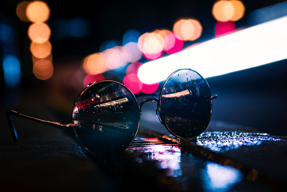 Обои для рабочего стола Очки в каплях дождя лежат на мокрой лавочке с отражением улицы ночного города