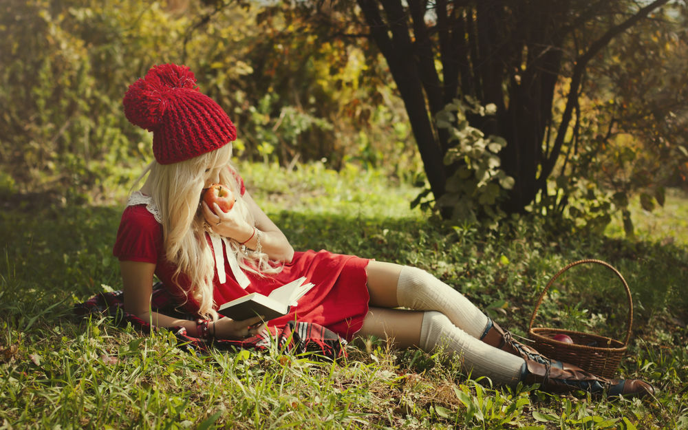 Обои для рабочего стола Девушка в красном платье и красной шапке лежит на траве возле дерева с яблоком в руке и читает книжку