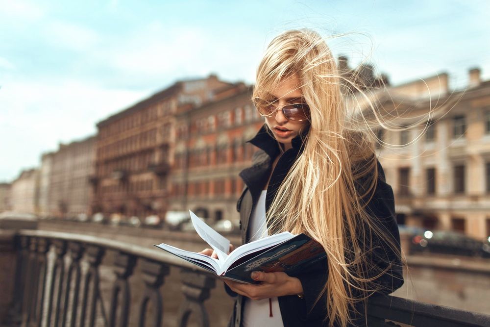 Обои для рабочего стола Длинноволосая блондинка с книгой в руках стоит у ограждения на набережной реки