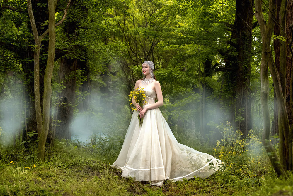 Обои для рабочего стола Девушка в длинном белом платье с цветами в руках стоит на фоне деревьев в тумане