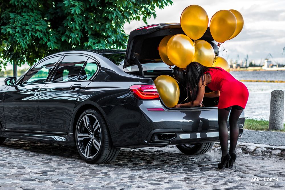 Обои для рабочего стола Девушка в красном платье и в черных чулках наклонилась к открытому багажнику авто BMW, фотограф Arny North