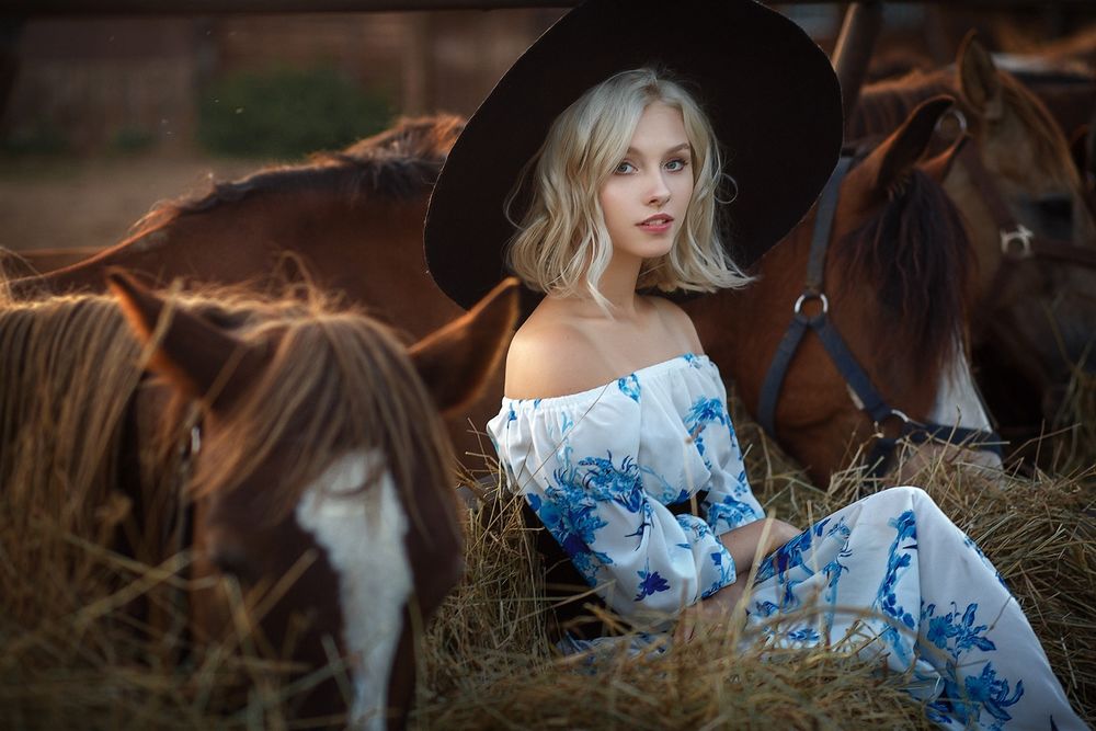 Обои для рабочего стола Модель Алиса Тарасенко в черной шляпе и в светлом цветастом платье сидит на сене между стоящими рядом лошадями, фотограф Настасья Паршина