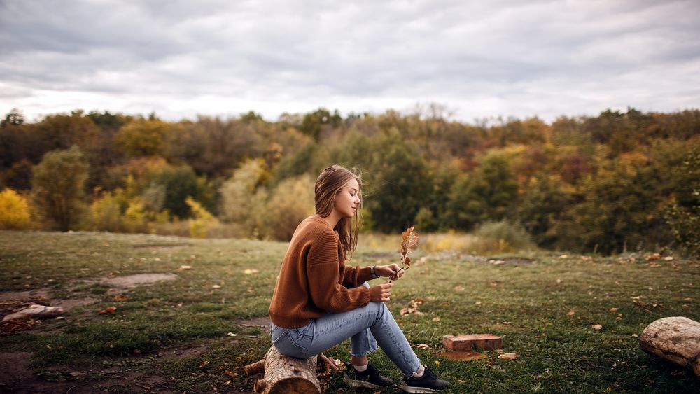 Обои для рабочего стола Девушка с осенним листиком в руке сидит на камне на фоне природы