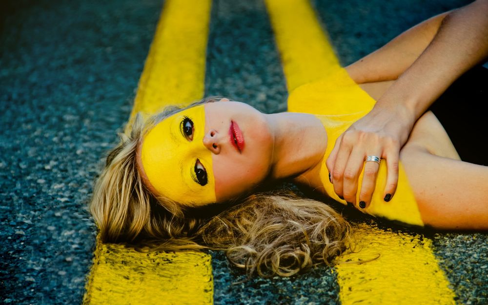 Обои для рабочего стола Портрет девушки лежащей на желтой двойной разделительной линии на дороге, на девушке краска повторяющая эти линии
