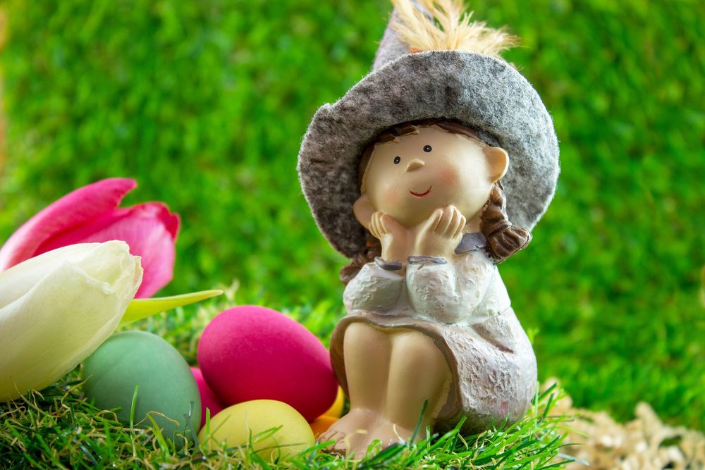 Обои для рабочего стола Игрушечная фигурка девочки в шляпе сидящая на траве на фоне пасхальных яиц и белого и розового тюльпанов, by Ingrid