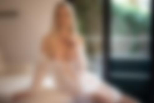 Обои для рабочего стола Блондинка Катя Енокаева с обнаженной грудью в приспущенном белом пеньюаре сидит на кровати в комнате на фоне окна