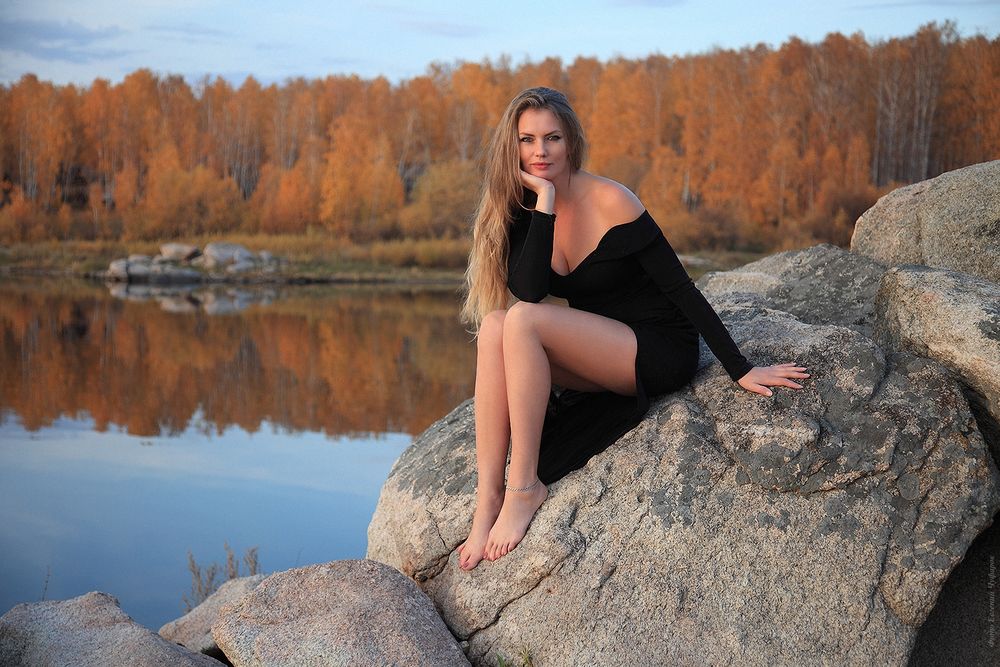 Обои для рабочего стола Длинноволосая блондинка - модель Ирина Никишина в черном платье сидит на камне на фоне водоема и осенней природы, фотограф Евгений Чуфаров