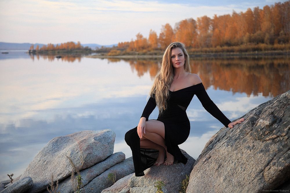 Обои для рабочего стола Длинноволосая блондинка - модель Ирина Никишина в черном платье сидит на корточках на камне на фоне водоема и осенней природы, фотограф Евгений Чуфаров