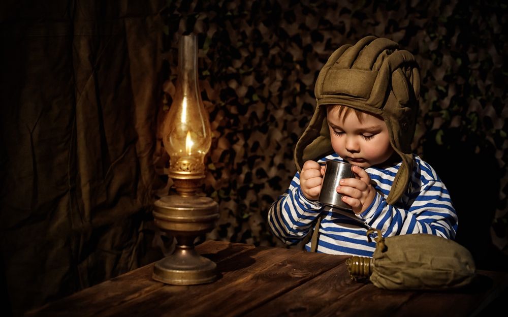 Обои для рабочего стола Мальчик в танкиском шлеме сидит за столом и при свете лампы пьет из кружки, фотограф Вера Калинкина