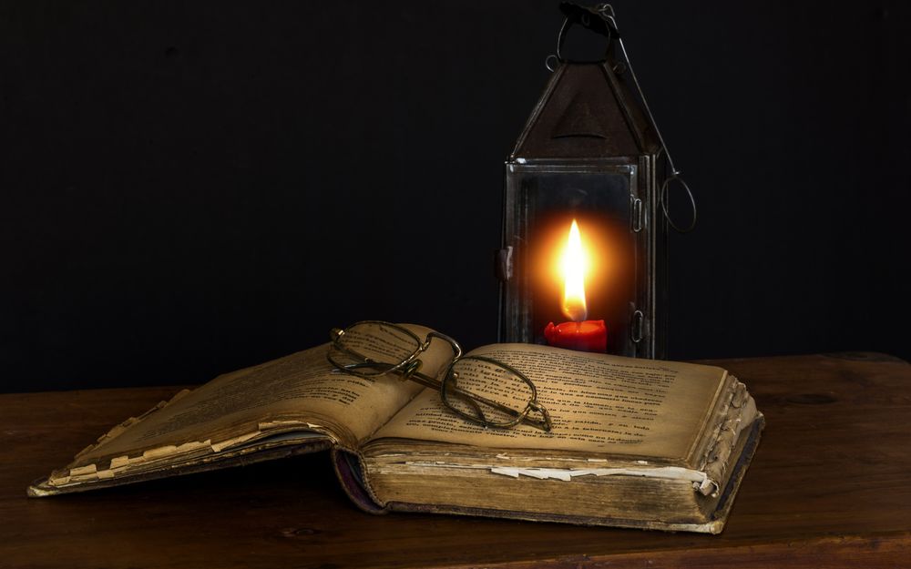 Обои для рабочего стола Раскрытая старая книга с очками лежат на столе на фоне фонаря со свечкой