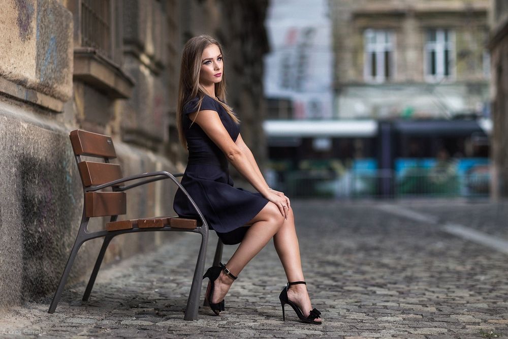 Обои для рабочего стола Модель Aлександра в черном платье сидит на скамейке у стены городского здания, фотограф Andrea Carretta