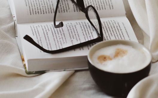 Обои на рабочий стол Чашка кофе с пенкой стоит на постели рядом с книгой, на которой лежат очки, автор Taisiia Shestopal