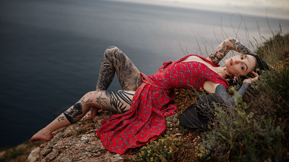 Обои для рабочего стола Модель Тина Рузина в красном платье с тату на теле лежит на вершине скалы морского берега, фотограф Георгий Чернядьев
