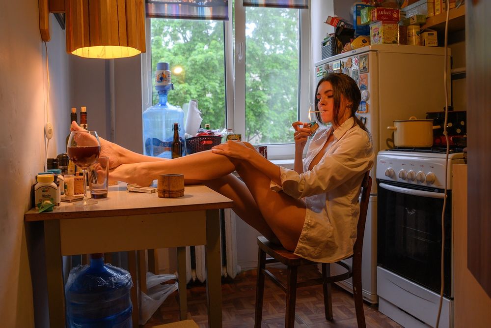 Обои для рабочего стола Модель Кристишка Шабанова в расстегнутой белой рубашке прикуривает сигарету, сидя на стуле на кухне, положив ноги на стол, фотограф Максим Лаухин