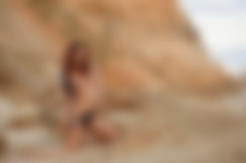 Обои для рабочего стола Рыжеволосая девушка Lelya Mult в приспущенных черных трусиках и с тату на ноге топлес стоит на коленях на песке на размытом фоне холма и неба, фотограф Николай Евдокимов