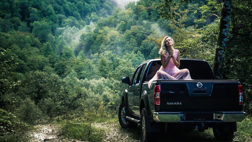 Обои для рабочего стола Блондинка в розовом платье сидит по-турецки на авто Nissan Navara на фоне природы