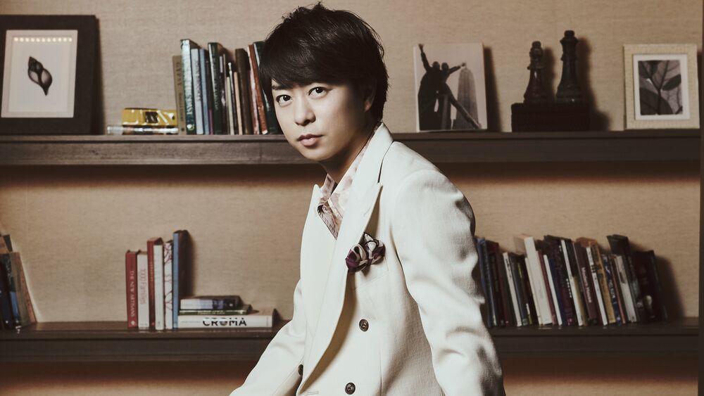 Обои для рабочего стола Певец, актер и телеведущий Сакурай Шо / Sakurai Sho из группы Араши / Arashi стоит в костюме на фоне полок, на которых есть книги, фото, картины и шахматы