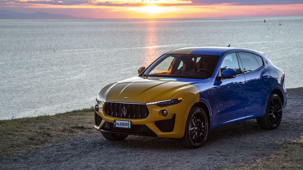 Обои для рабочего стола Суперкар желто-голубого цвета Maserati Levante GT Hybrid стоит возле озера на фоне заката солнца