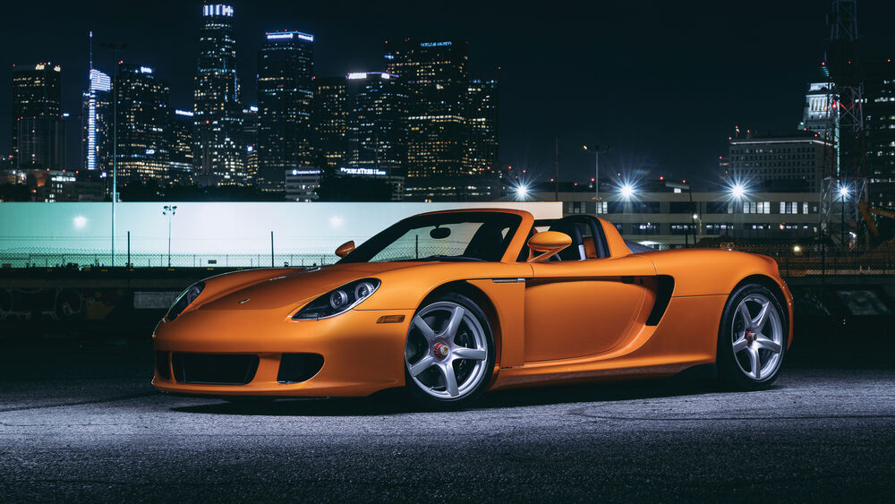 Обои для рабочего стола Суперкар оранжевого цвета Порше / Porsche Carrera GT стоит на фоне ночного города