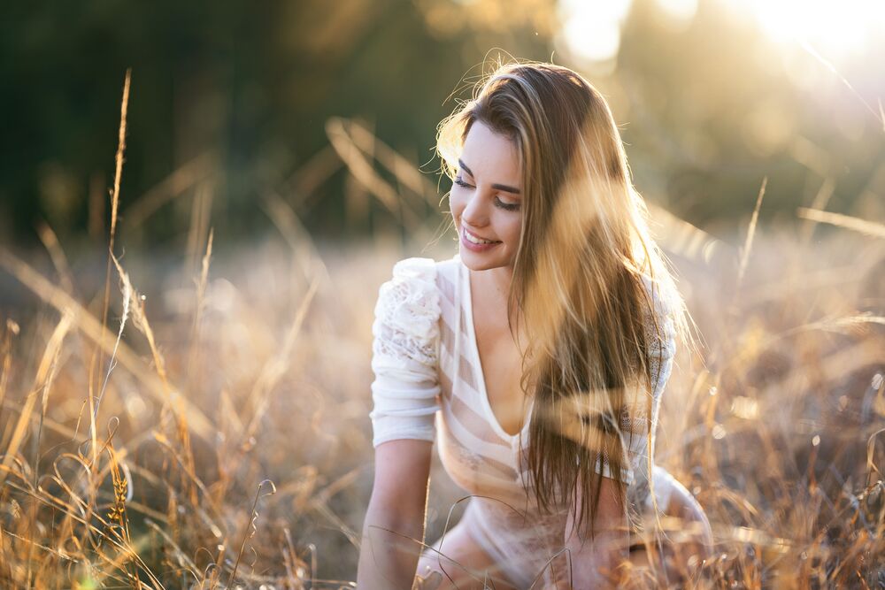 Обои для рабочего стола Светловолосая девушка - модель Alexis Rose с белом платье и с улыбкой на лице сидит в поле с травой на размытом фоне природы, фотограф Christopher Rankin