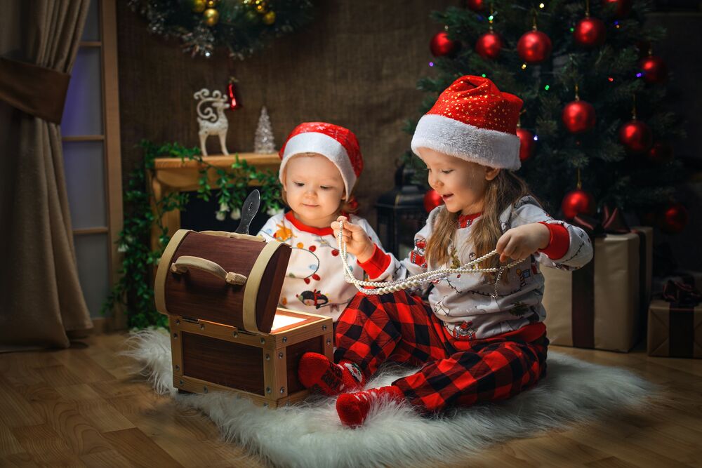 Обои для рабочего стола Маленькие дети в новогодних колпаках сидят на полу у открытого сундучка в комнате с новогодней елкой