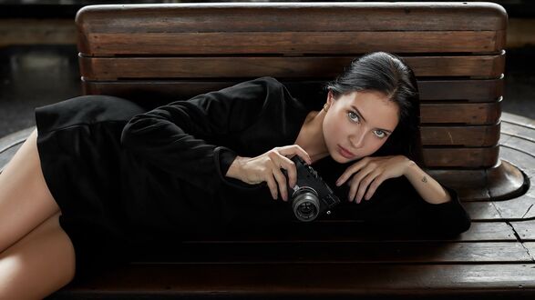 Обои на рабочий стол Модель Aigul Fazylova в черном платье с фотоаппаратом в руке лежит на деревянной лавочке