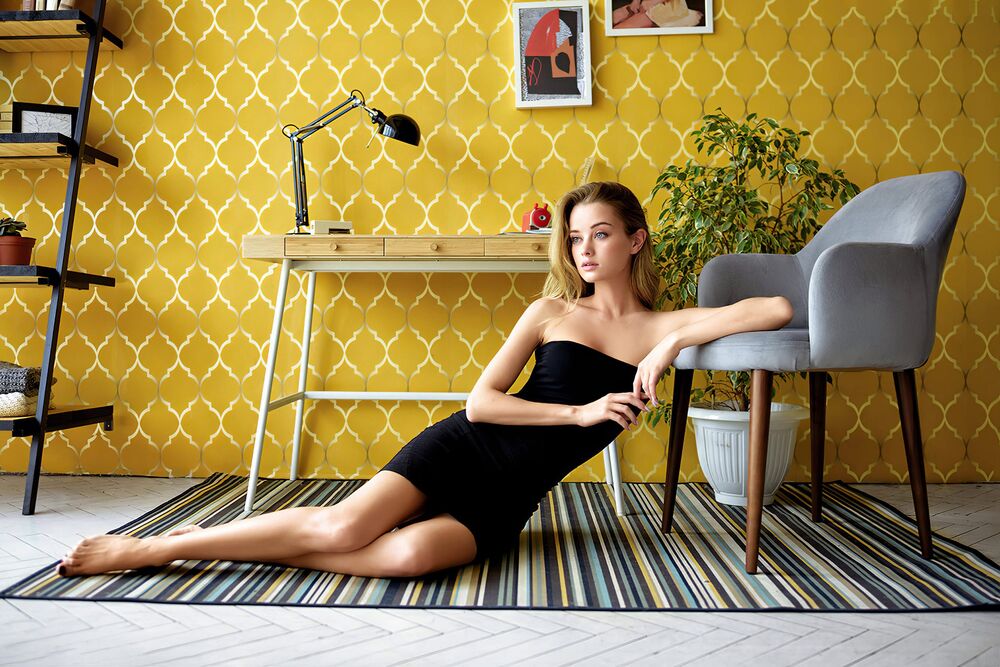 Обои для рабочего стола Модель Мария Жгенти в черном платье сидит на коврике на полу комнаты, облокотившись на кресло