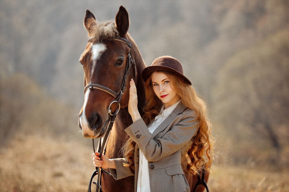 Обои для рабочего стола Рыжеволосая девушка в шляпе и в пиджаке стоит рядом с лошадью на размытом фоне природы