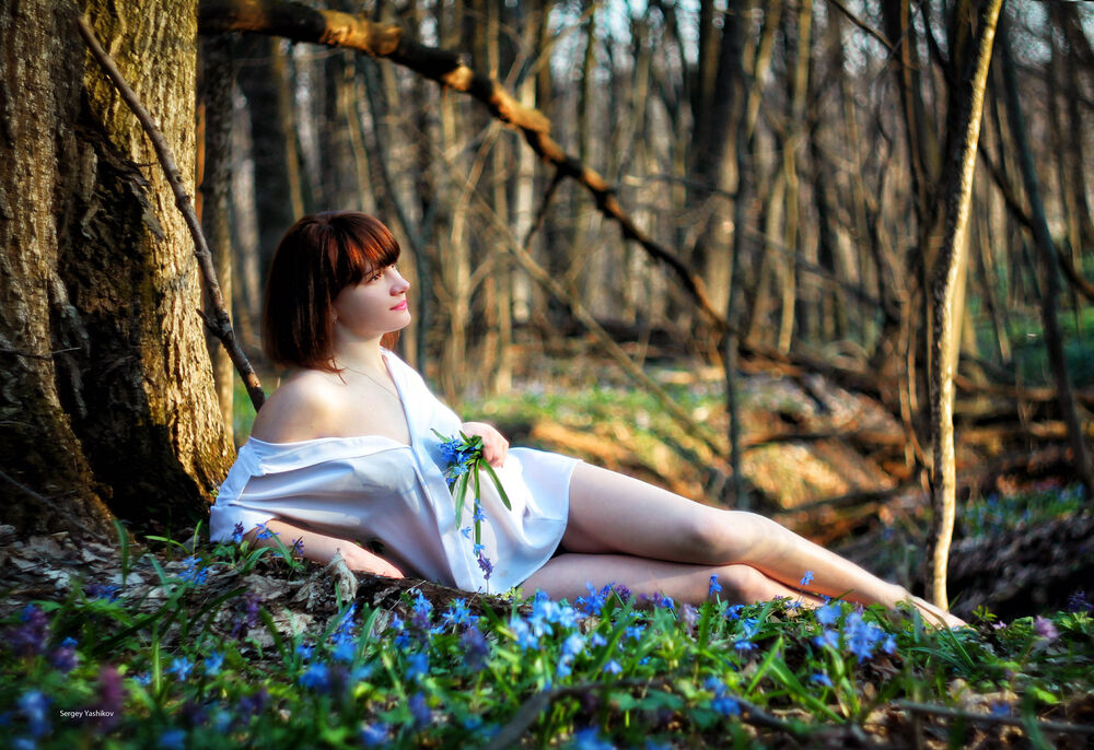Обои для рабочего стола Шатенка в белой рубашке лежит у дерева в весеннем лесу и держит в руке цветы подснежники