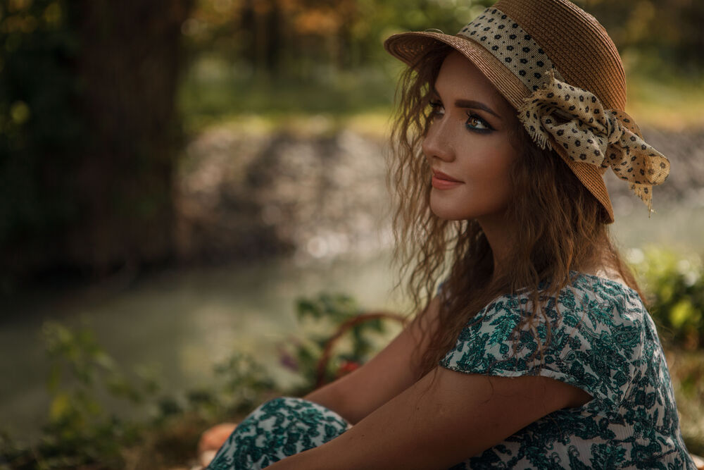 Обои для рабочего стола Девушка в летнем платье и шляпке сидит у реки