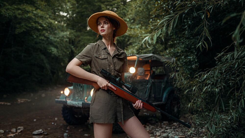 Обои для рабочего стола Модель Виктория Агеева в шляпе и комбинезоне цвета хаки со снайперской винтовкой в руках стоит на дороге поодаль от авто