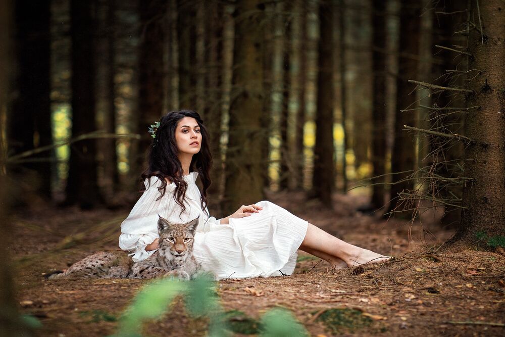 Обои для рабочего стола Модель Алена Беляева в белом платье сидит в лесу с лежащей рядом рысью