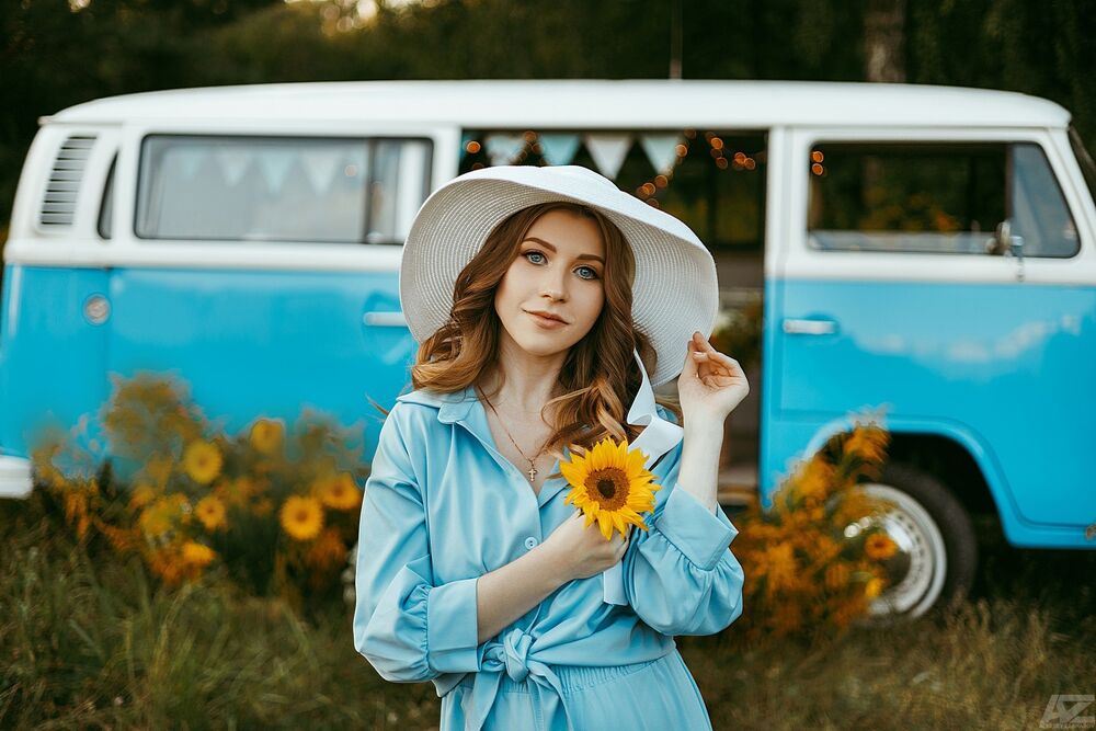 Обои для рабочего стола Модель Валерия Козырева в шляпе и в голубом платье стоит на природе на фоне авто с цветком в руке