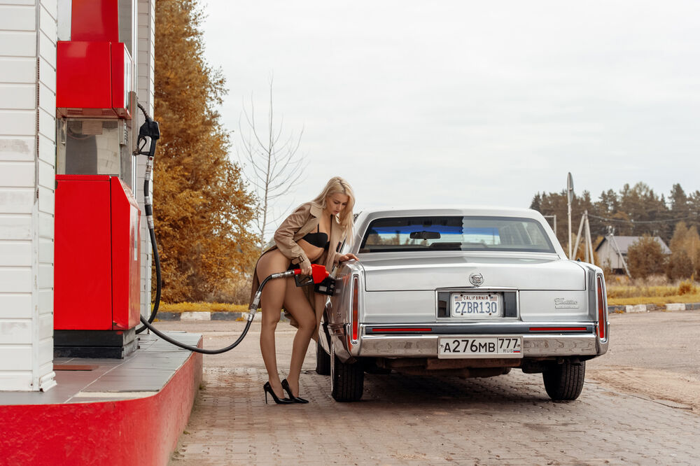 Обои для рабочего стола Блондинка в расстегнутом плаще и в нижнем белье стоит на АЗС и заправляет авто Cadillac De Ville
