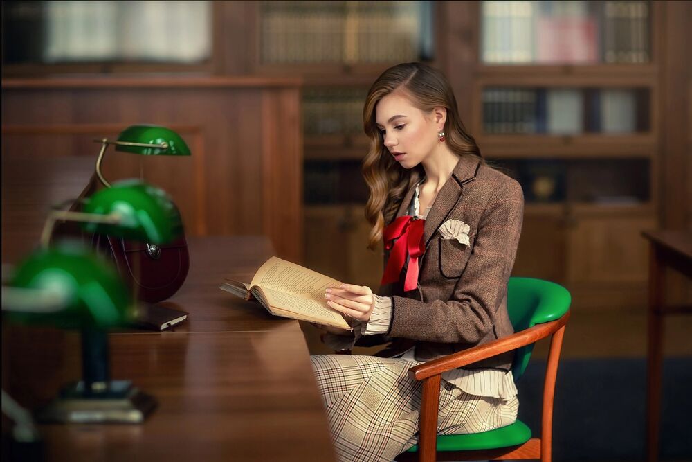Обои для рабочего стола Длинноволосая девушка в пиджаке с бантом и в сарафане сидит на кресле за столом с книгой в руках в читальном зале